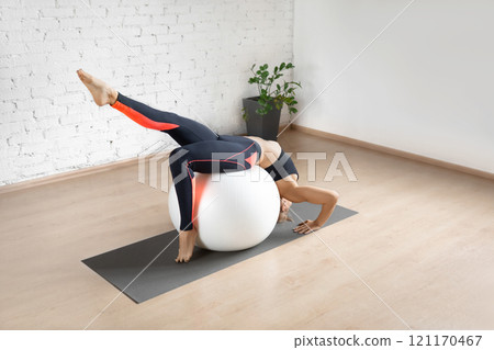Fit woman in black sportswear does gymnastic bridge exercise with one leg up using fitness ball in loft studio indoor. 121170467