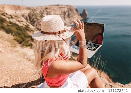 A woman is sitting on a rocky beach with a laptop in front of her. She is wearing a pink top and a straw hat. Concept of relaxation and leisure. 121172741