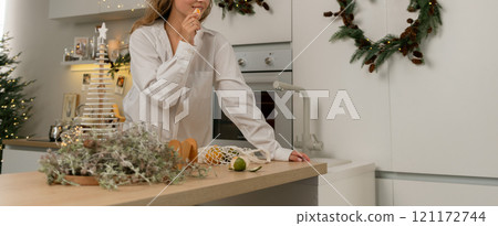 A woman is standing in a kitchen with a Christmas tree in the background. She is wearing a white robe and is preparing food on a counter. A woman is standing in a kitchen with a Christmas tree in the background. She is wearing a white robe and is preparing food on a counter. 121172744