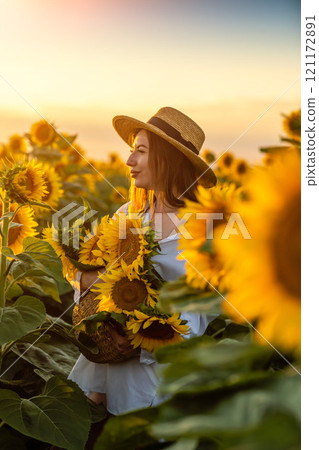 A girl in a hat on a beautiful field of sunflowers against the sky in the evening light of a summer sunset. Sunbeams through the flower field. Natural background. 121172891