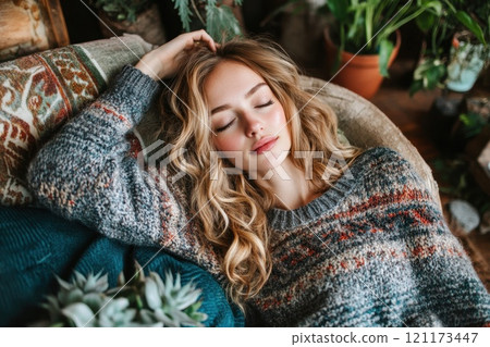 A young woman with wavy blonde hair performs EFT tapping (Emotional Freedom Technique) on her head while resting on a cozy couch, surrounded by blankets and indoor plants 121173447