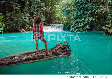 Woman in a shirt standing on a log over a turquoise river in the jungle with a waterfall. Tropical adventure. 121173979