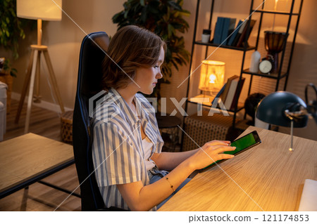 A Young Woman Engaged in Working Late into the Evening From the Comfort of Her Home Using a Tablet 121174853
