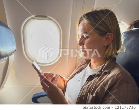 Passenger woman is flying in plane. Girl using phone sitting by airplane window. Traveling female with smartphone inside plane enjoying flight. Woman using wifi connection on board. Traveling girl Passenger woman is flying in plane. Girl using phone sitting by airplane window. Traveling female with smartphone inside plane enjoying flight. Woman using wifi connection on board. Traveling girl 121175736