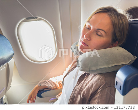 Rest on plane. Young woman napping sitting by porthole in business jet using neck pillow for travel. Passenger girl flying in plane. Girl sleeping in airplane going on trip vacation. Traveling girl Rest on plane. Young woman napping sitting by porthole in business jet using neck pillow for travel. Passenger girl flying in plane. Girl sleeping in airplane going on trip vacation. Traveling girl 121175738