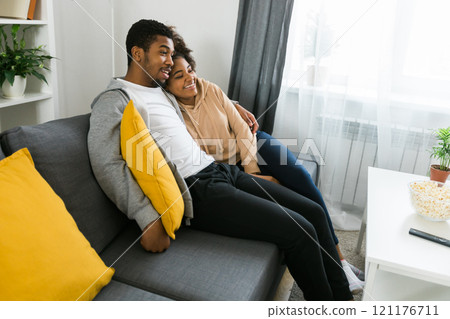 African American couple relaxes on a cozy sofa, sharing a joyful moment while watching a show. Essence of quality time and comfort in a stylish modern living room African American couple relaxes on a cozy sofa, sharing a joyful moment while watching a show. Essence of quality time and comfort in a stylish modern living room 121176711