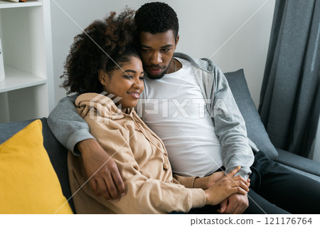 African American couple sits on a cozy gray sofa, embracing each other and enjoying a peaceful moment at home. Love and intimacy and a sense of comfort in a modern and inviting interior 121176764