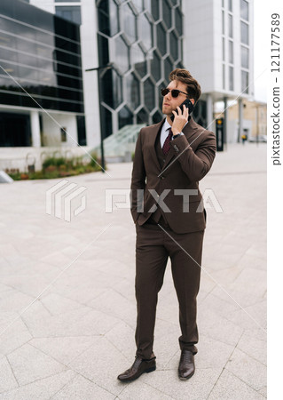 Full length vertical portrait of successful businessman in suit and sunglasses making call using mobile phone standing in city street on cloudy day. Concept of business communication and technology. 121177589