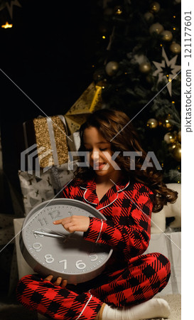 Little girl in night pajamas holding a watch in her hands waiting for Santa Claus and magic. Christmas night, festive atmosphere. Little girl in night pajamas holding a watch in her hands waiting for Santa Claus and magic. Christmas night, festive atmosphere. 121177601