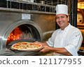 The chef Man smiling at a table with pizza, plant, and cheese in front of him 121177955