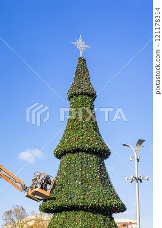Large outdoor Christmas tree being decorated under a clear blue sky with star on top 121178314