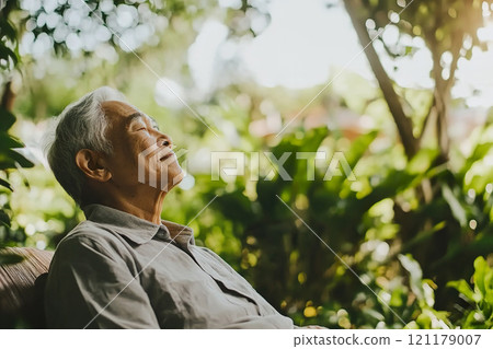 Elderly man relaxing outdoors in park surrounded by lush greenery during a sunny afternoon. 121179007