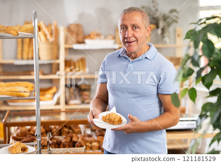 Senior male customer stands with croissant in hands near window of bakery. Senior male customer stands with croissant in hands near window of bakery. 121181529