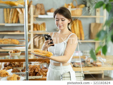 Beautiful young girl using smartphone choosing bocadillo in bakery shop Beautiful young girl using smartphone choosing bocadillo in bakery shop 121181624