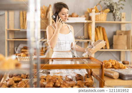 European girl in bakery is talking on phone, inspecting goods in window 121181952