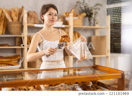 Beautiful girl shopper selecting and putting fresh baked croissants into plastic bags from shelf in bakery shop 121181969