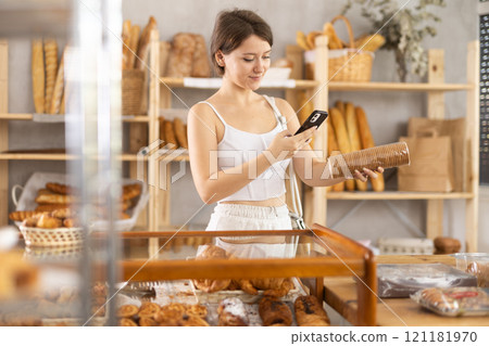 European young woman customer at bakery scans QR code on cookie package using phone camera 121181970