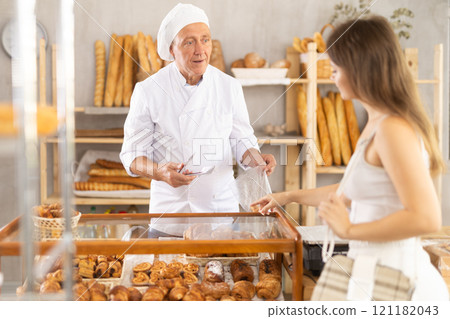 Beautiful interested young girl buyer points to products on display and chooses baked goods, elderly man seller helping her in bakery store Beautiful interested young girl buyer points to products on display and chooses baked goods, elderly man seller helping her in bakery store 121182043