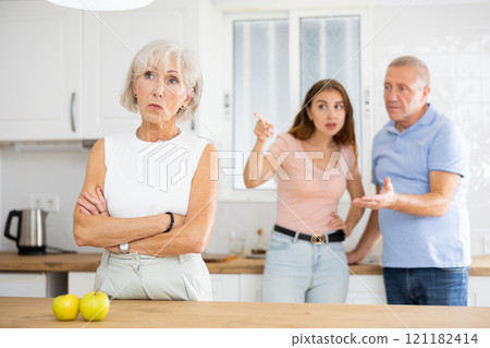 Elderly frustrated woman turned away from scolding her husband and adult daughter in kitchen Elderly frustrated woman turned away from scolding her husband and adult daughter in kitchen 121182414