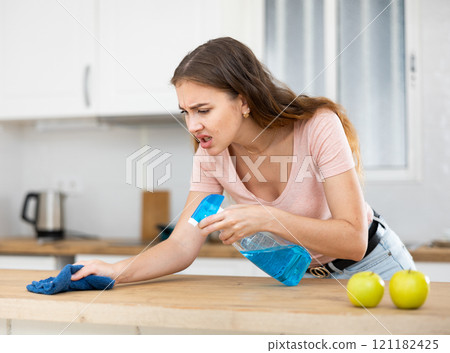Displeased housewife cleaning dinning table with rag in kitchen Displeased housewife cleaning dinning table with rag in kitchen 121182425
