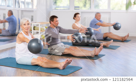 Group of mature people in sportswear exercising Pilates with soft balls while sitting on mats in fitness studio Group of mature people in sportswear exercising Pilates with soft balls while sitting on mats in fitness studio 121182564
