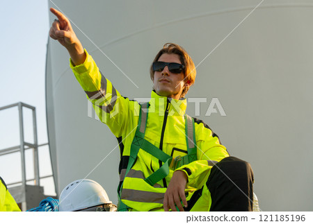 Portrait of wind turbine worker or technician man sit on base with point to his right side with pole of wind turbine in the background. 121185196