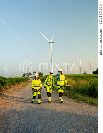 Vertical image of group of wind turbine or windmill workers or technicians carry tools and walk along the road in workplace field with row of wind turbine in the background. 121185199
