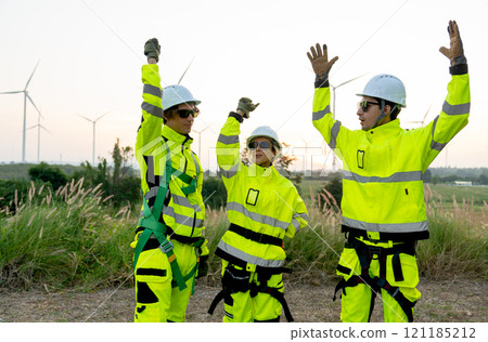 Group of wind turbine workers or technicians raise their hands up together with warm light and row of wind turbine in the background. 121185212