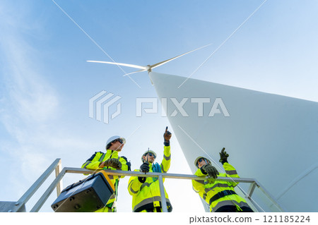 Wide shot and lower view of group of wind turbine or windmill workers or engineers discuss together on base of pole of wind turbine in workplace field. 121185224