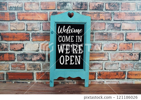 Welcome We're Open text on blackboard easel stand on wooden and old brick wall background 121186026