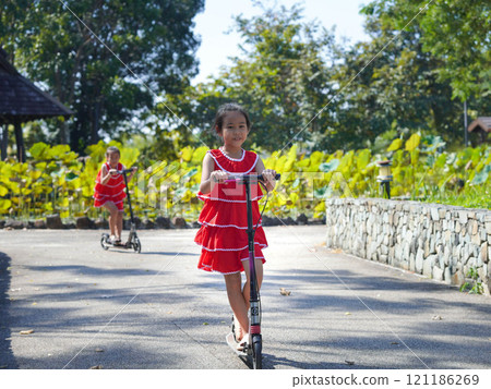 Cute sisters riding scooters on a sunny summer day in the park. Active leisure and outdoor sport for child. Cute sisters riding scooters on a sunny summer day in the park. Active leisure and outdoor sport for child. 121186269