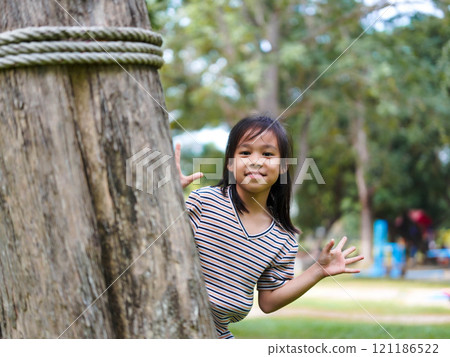 Cute little girl is peeking behind a tree, having fun playing playing peek-a-boo in a sunny garden. Smiling child in the park 121186522