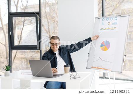 A man dressed in casual clothes works in the office, sits at a table near a laptop and holds an online conference. 121190231