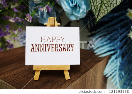 Happy Anniversary text write on paper card with wooden easel and flowers decoration on wooden background 121193388