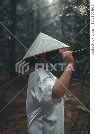 A young man in a white shirt and a conical hat stands in the rain holding a katana sword on his shoulder. The mysterious atmosphere reminds of a warrior or samurai in a mystical forest with a katana. 121195608