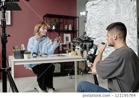 Male operator kneeling holding camera and shooting actress who sitting at table with cosmetics at studio Male operator kneeling holding camera and shooting actress who sitting at table with cosmetics at studio 121195764