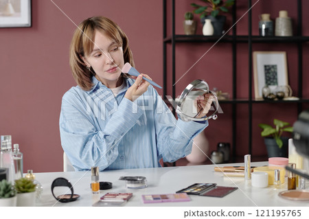 Relaxed woman holding mirror while looking in it and applying blush cheeks Relaxed woman holding mirror while looking in it and applying blush cheeks 121195765