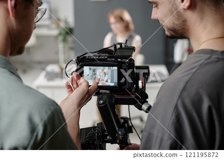 Medium close up of unrecognizable videographers looking at camera display and discussing scene with model who sitting at table in background 121195872