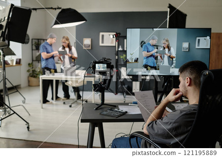 Unrecognizable man sitting at table and reading script while his coworkers discussing props and decorations for shooting space in background Unrecognizable man sitting at table and reading script while his coworkers discussing props and decorations for shooting space in background 121195889