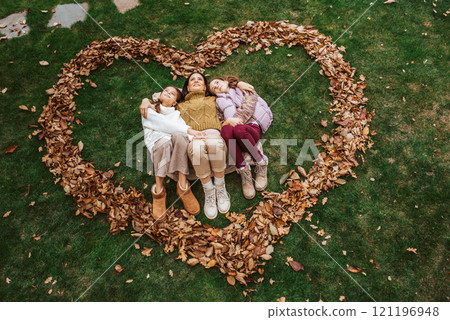 Portrait of mother with two daughters, laying on grass in the middle of heart from leaves. 121196948
