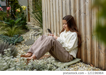 Young girl sitting on ground outdoors, holding smartphone and scrolling. Young girl sitting on ground outdoors, holding smartphone and scrolling. 121196964