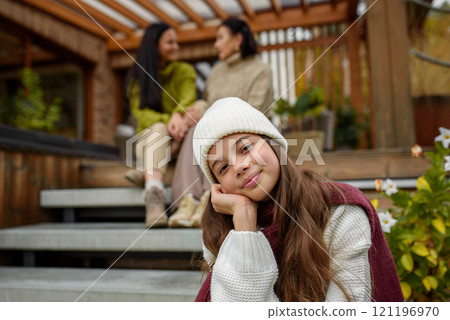 Portrait of young girl sitting on wooden stairs, mom and grandmother behing her. Mother's day and multigenerational family concept. 121196970