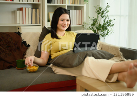 Relaxed young woman watching videos on digital tablet and enjoying snacks Relaxed young woman watching videos on digital tablet and enjoying snacks 121196999