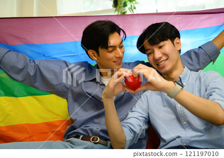 Smiling young men couple holding a red heart and sitting under rainbow pride flag 121197010