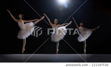 Three young girls, ballet dancers in classical white tutus performing graceful dance under spotlight. Beautiful and artistic ballet performance 121197536