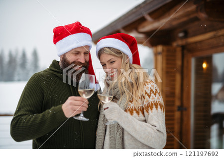 Mature couple wearing Santa hats, standing in front cabin and clinking with wine glasses, toast. 121197692