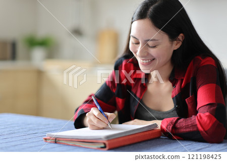 Happy asian student writing notes on notebook at home 121198425