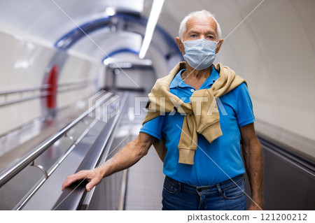 Elderly man in protective mask moving up on escalator while leaving subway station platform 121200212