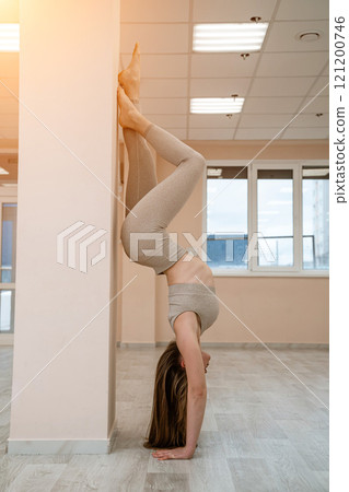 The girl is training in the yoga studio. Dressed in a beige tracksuit doing a handstand against the wall. Healthy lifestyle and yoga concept. 121200746