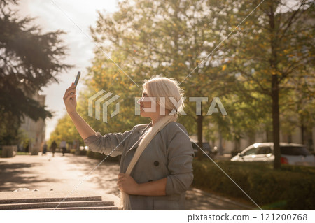 A woman is taking a picture of herself with her cell phone. She is wearing a gray jacket and scarf. The scene is set in a city with trees and cars in the background. 121200768